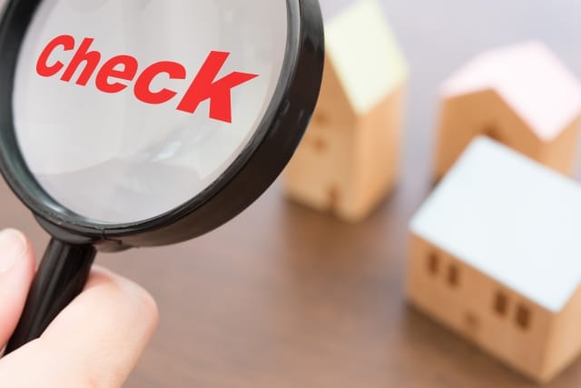 木製の家の模型を背景に、虫眼鏡で「Check」という赤い文字を拡大している