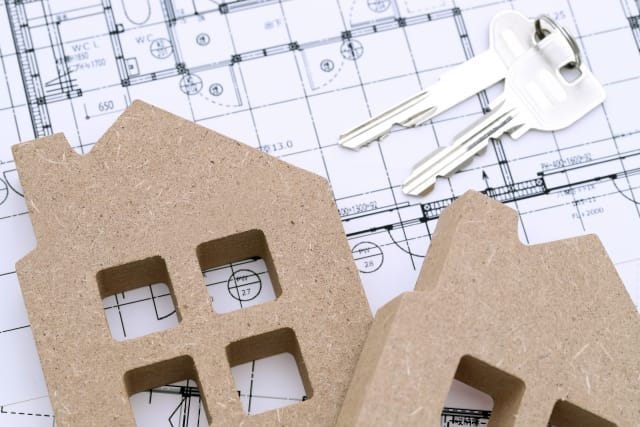住宅の設計図の上に、家の形をした木製オブジェと銀色の鍵が置かれている