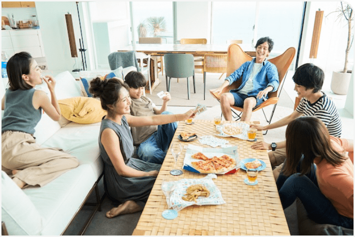 リビングルームで6人の男女がピザや軽食を囲んで楽しそうに過ごしている様子