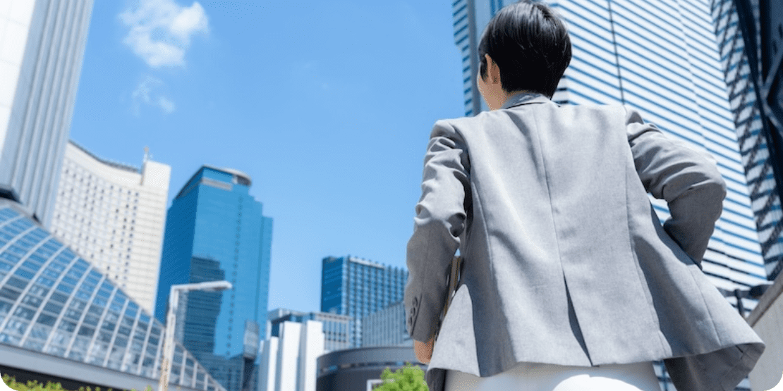 スーツ姿のビジネスパーソンが高層ビル群を見上げながら街中を歩いている
